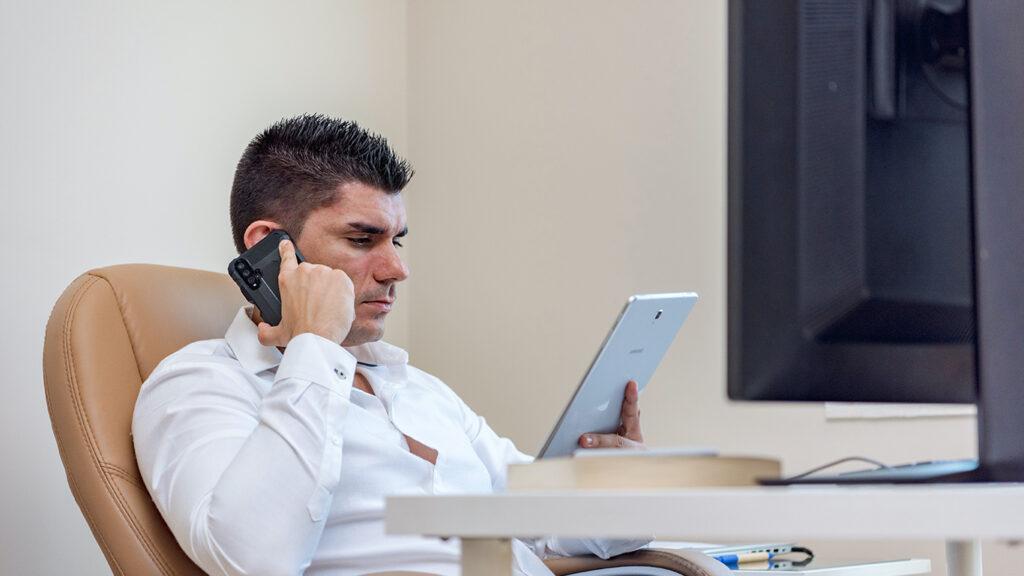 A Man in White Long Sleeve Shirt Using a Phone While Holding a Tablet
