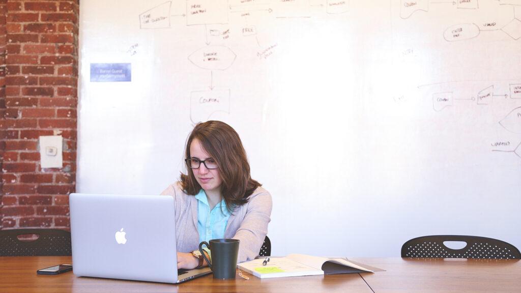 Woman Wearing Black Framed Eyeglasses and Teal Button-up Shirt and Beige Blazer Sitting at Table Near White Macbook