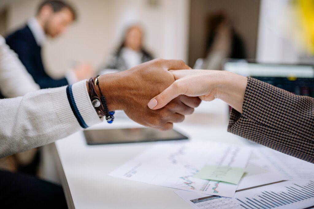 Two individuals engaging in a handshake over a business desk with documents, indicating a professional agreement