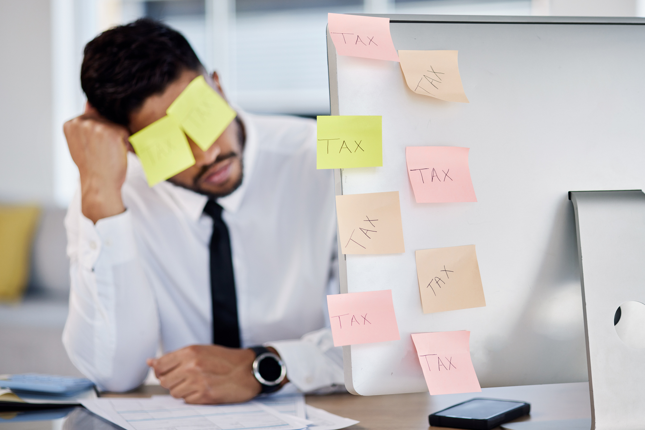 A man at a desk appears overwhelmed by tax work, with sticky notes labeled "TAX" on his forehead and computer